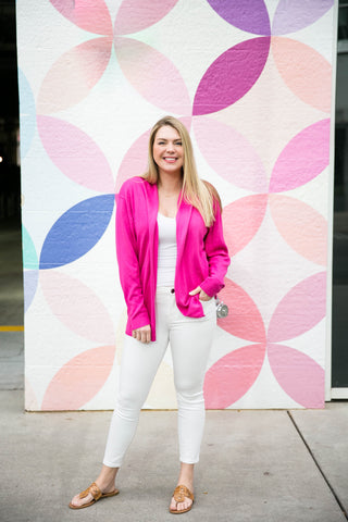Woman in a bright pink jacket and white pants standing in front of a colorful circular patterned wall.