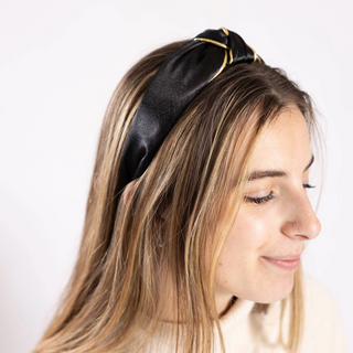 Model wearing black Lanie Headband – Woman in cream turtleneck wearing the Lanie Headband in black satin with gold trim and knotted detail.