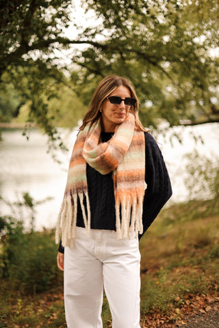 Lifestyle shot of Hollis Scarf in camel and green stripes styled outdoors with a navy sweater and white pants – trendy fall scarf look.
