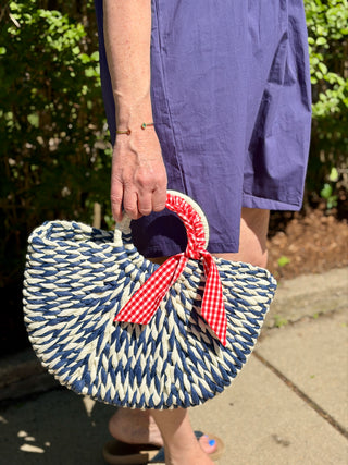 Person holding a blue and white straw bag with a red and white checkered ribbon outdoors.