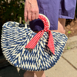Person holding a blue and white striped woven straw handbag accented with a red and white gingham ribbon.