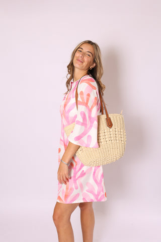 Woman wearing a pink and white abstract dress holding a cream straw bag 