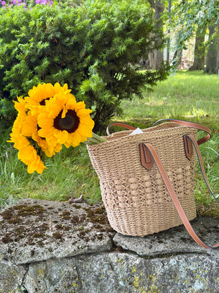 Natural woven handbag with a brown straps with fresh sunflowers propped up on a stone ledge outdoors.