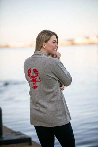 Woman wearing a gray jacket with a red lobster design by a body of water.