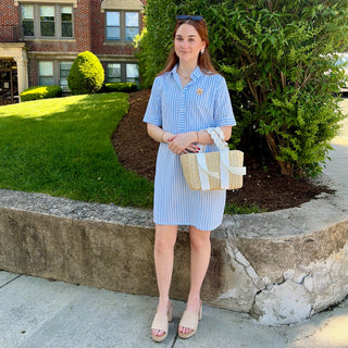 Woman in a blue and white striped dress standing outdoors holding a cream straw woven handbag with cream ribbon handles