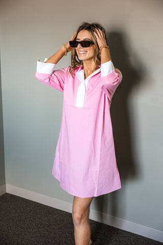 Woman wearing a pink dress with white collar and cuffs against a plain wall.