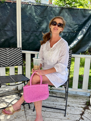 Woman sitting on a chair with a pink bag and iced drink, wearing sunglasses and a white outfit.