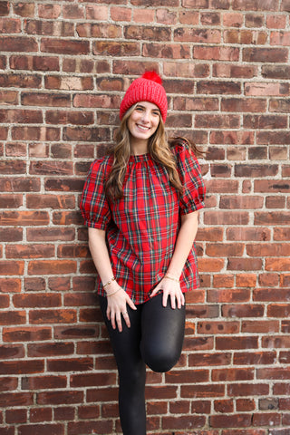 Woman wearing a red plaid top and red beanie against a brick wall