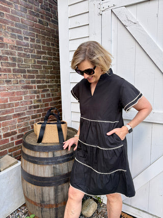 Woman in a black dress standing next to a wooden barrel with a natural straw woven handbag with black ribbon handles propped up on it.