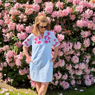 Woman in a light blue dress and white striped with pink floral patterns standing in front of a bush of pink flowers.