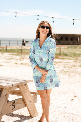 Woman in a patterned dress standing on a beach with a wooden picnic table and ocean view.