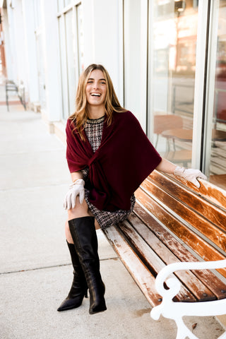 Woman sitting on a bench wearing a burgundy shawl and knee-high boots, smiling.