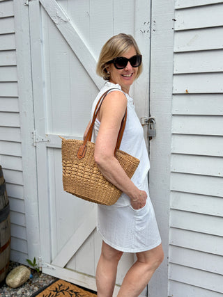 Woman in a white dress holding a natural woven handbag, standing in front of a white wooden door.