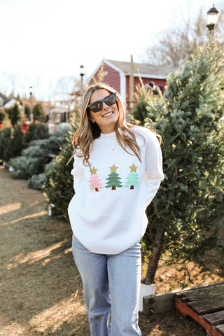 Woman wearing Pippa White Christmas Sweater with embroidered holiday trees topped with gold stars, styled with jeans outdoors.
