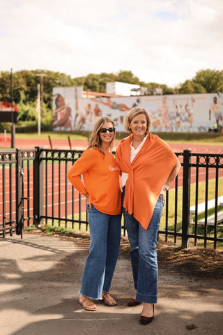 Two women in orange shirts standing on a track field with a mural in the background.