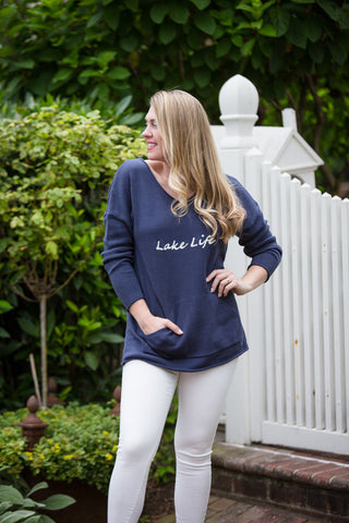 Woman wearing a navy blue 'Lake Life' sweatshirt and white pants standing outdoors.