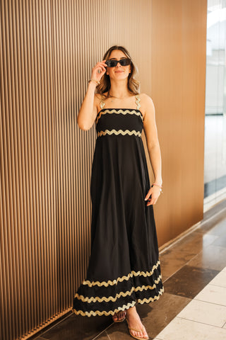 Woman in a black dress with gold trim standing against a textured wall.