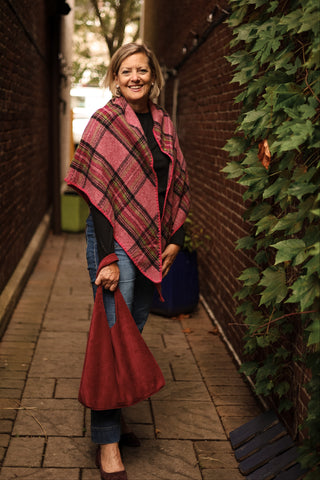 Woman wearing a pink plaid shawl and holding a bordeaux hobo bag in an alleyway.
