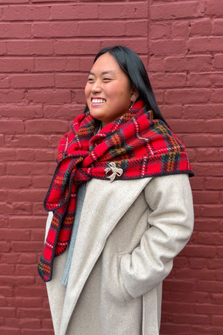 Person wearing a red plaid scarf and beige coat against a brick wall.
