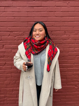 Woman wearing a red plaid scarf and beige coat holding a glass of red wine against a red brick wall.