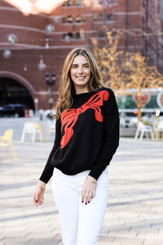Black Sweater Image: Woman wearing black Madeline Holiday Bow Sweater with red bow detail – cozy and stylish holiday outfit.