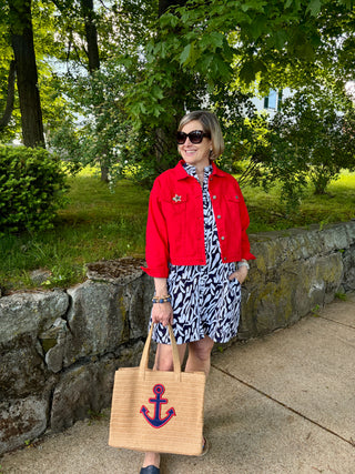 Woman in a red jacket and patterned dress holding a tote bag with an red and navy anchor design, standing outdoors.