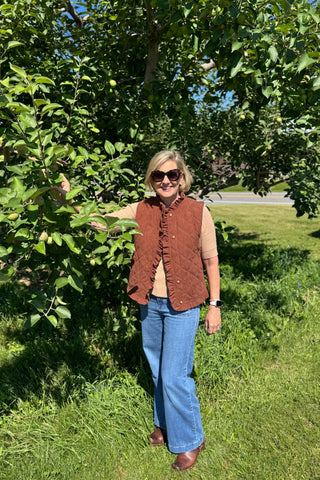 A woman wearing a brown quilted sleeveless vest over a beige top, blue jeans, and brown shoes stands outdoors in a grassy area next to a leafy tree.