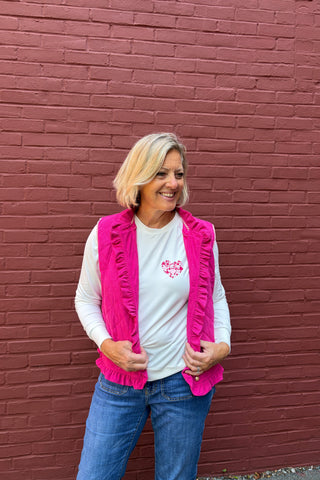 A woman with blonde hair stands in front of a red brick wall wearing a white long-sleeve t-shirt with a pink heart graphic, a bright pink ruffled vest, and blue jeans.