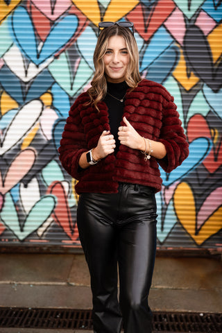 Woman wearing Callie Bolero Vest in Bordeaux wine – cropped faux fur jacket styled with black turtleneck and leather pants.