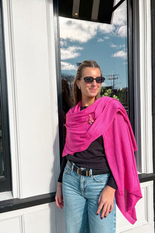 Woman wearing a bright pink scarf, black sunglasses, and blue jeans standing indoors with a window in the background.