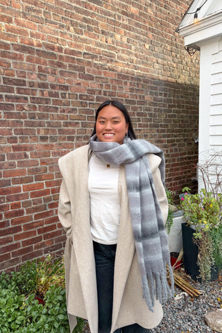 Person wearing a beige coat and gray scarf standing in front of a brick wall.