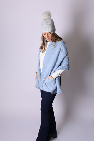 Woman wearing silver Chrissy ribbed knit hat with faux fur pom pom, with a light blue wrap and blue pants.