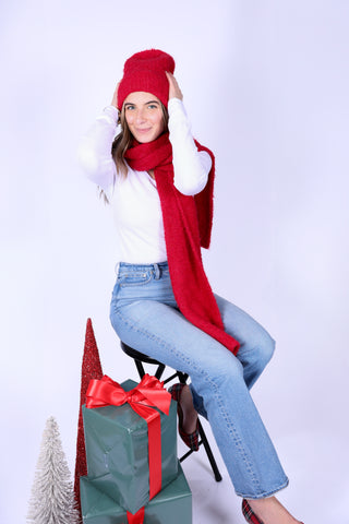Woman wearing a red beanie and scarf, sitting on a stool with a large gift box and tree against a white background