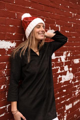 Woman smiling in festive red and white holiday beanie with pom pom against brick wall