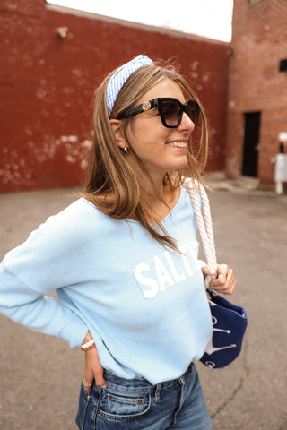Woman wearing a light blue sweatshirt with text, sunglasses, and a headband outdoors.