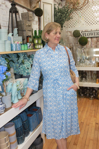 Woman wearing a blue patterned dress in a store setting with floral arrangements and shelves.