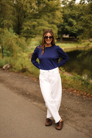Woman in a navy bubble sleeve tshirt top and white pants standing on a path with trees and water in the background