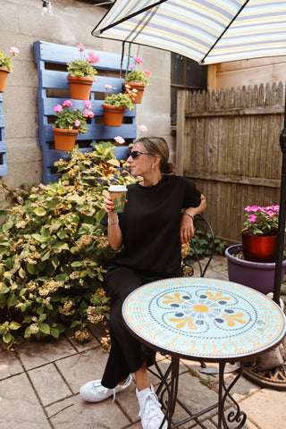 Woman sitting at a table outdoors with potted plants and a striped umbrella.
