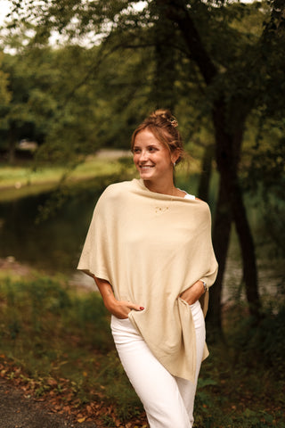 Woman in a beige poncho and white pants standing outdoors with trees and water in the background
