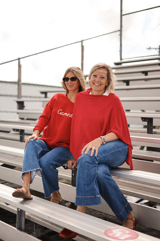 Two women sitting on bleachers wearing red sweatshirts with text.