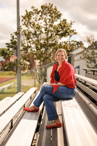 Woman sitting on bleachers at a sports field wearing a red shirt and blue jeans.