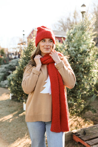 Woman wearing a red beanie and scarf with a beige sweater featuring a Christmas tree design, standing in a Christmas tree lot.