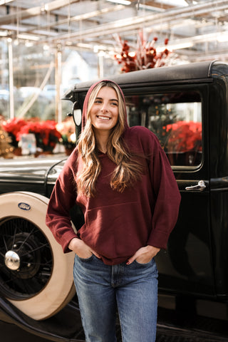 Woman in a maroon sweatshirt standing next to an old car in a greenhouse setting
