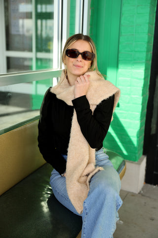 Woman sitting on a bench wearing sunglasses and a black and beige cardigan against a green wall.