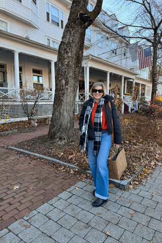 Woman standing on a sidewalk in front of a house with an American flag, wearing a black coat and blue jeans.